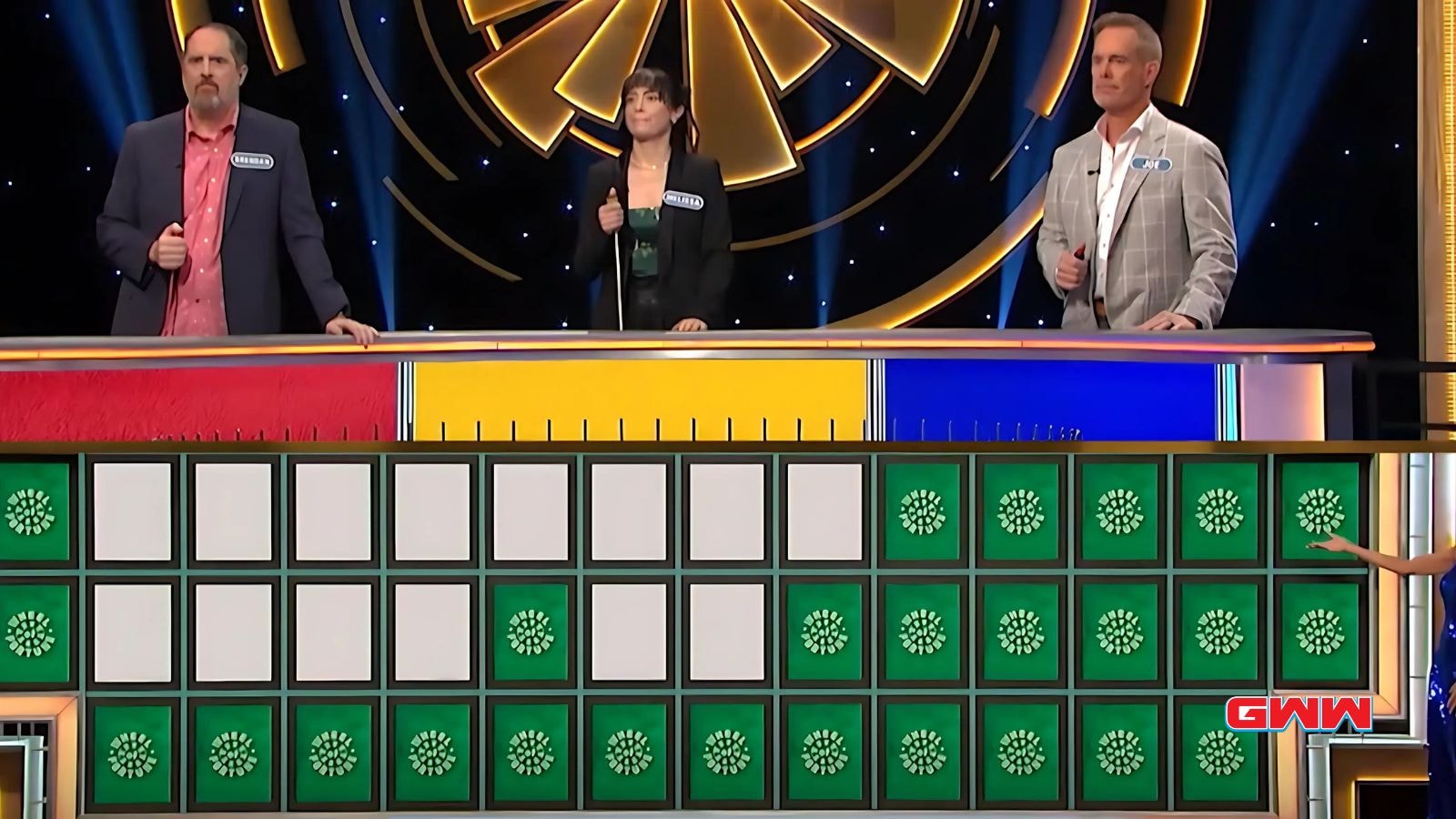 Game show contestants Brendan, Melissa, and Joe stand behind a puzzle board fortune celebrity wheel.