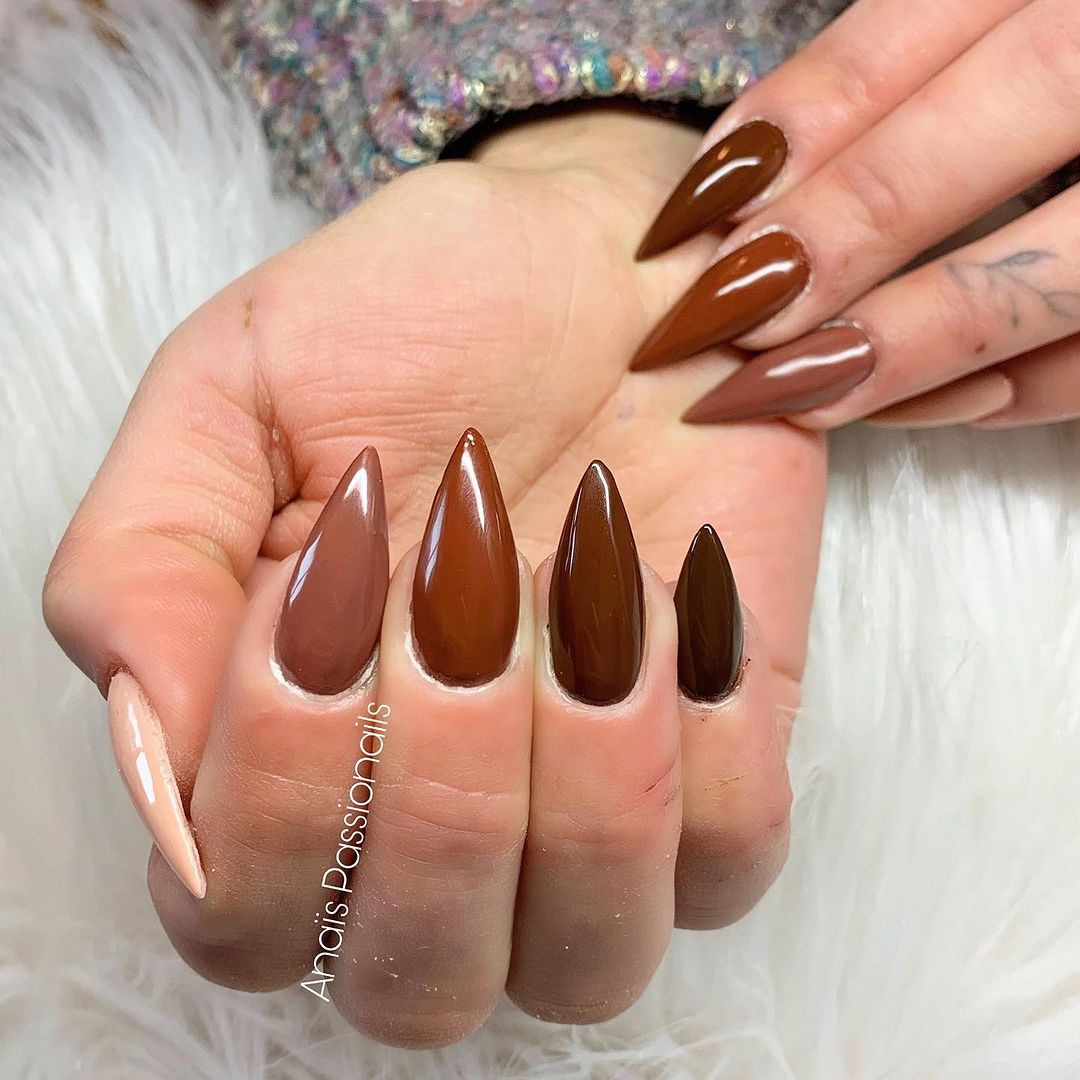 A hand with long, stiletto-shaped nails in a gradient of brown shades against a white furry background.

