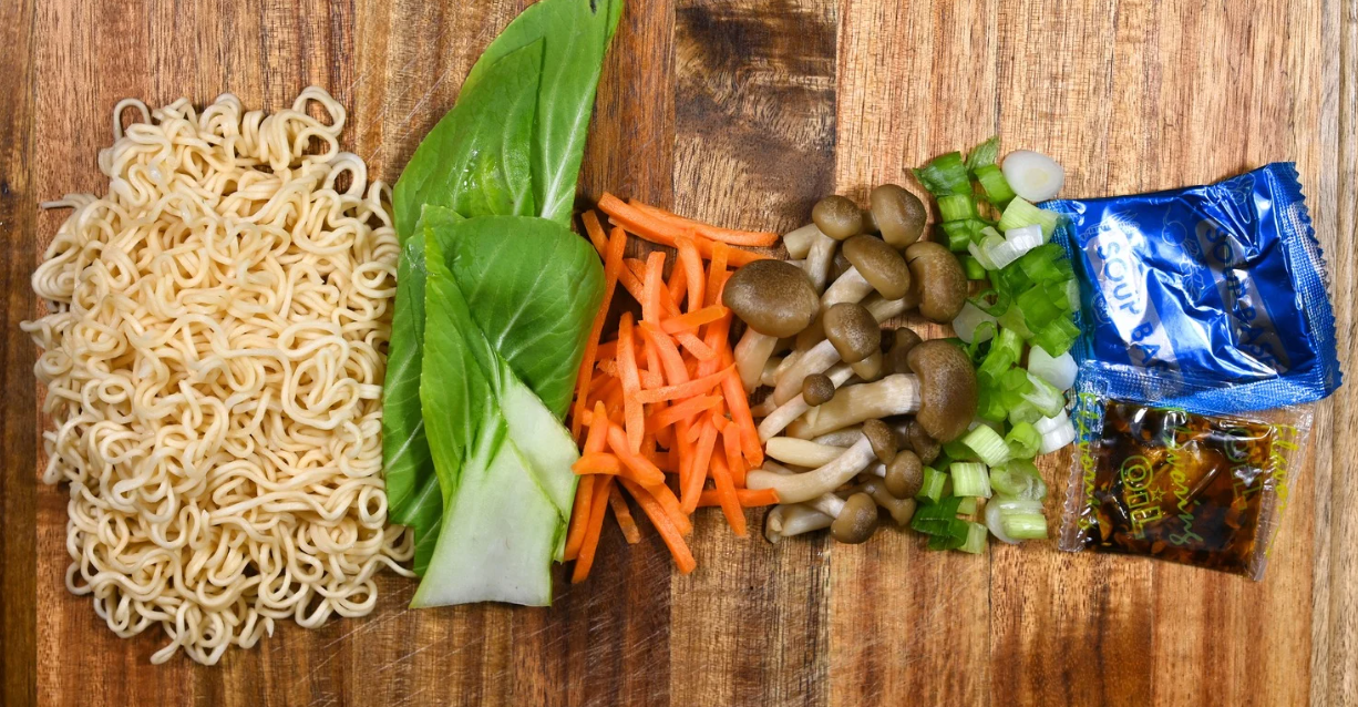 (Flache Lage ungekochter Instantnudeln, Pak Choi, in Julienne geschnittene Karotten, Pilze, gehackte Frühlingszwiebeln und zwei Gewürzpakete auf einem Holzschneidebrett.)