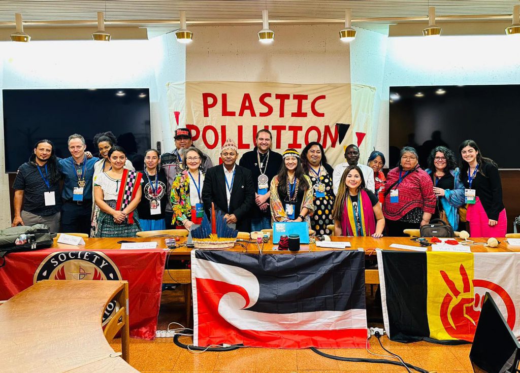 A group of diverse people behind a long table with flags, one the tino rangatiratanga flag. A banner says PLASTIC POLLUTION