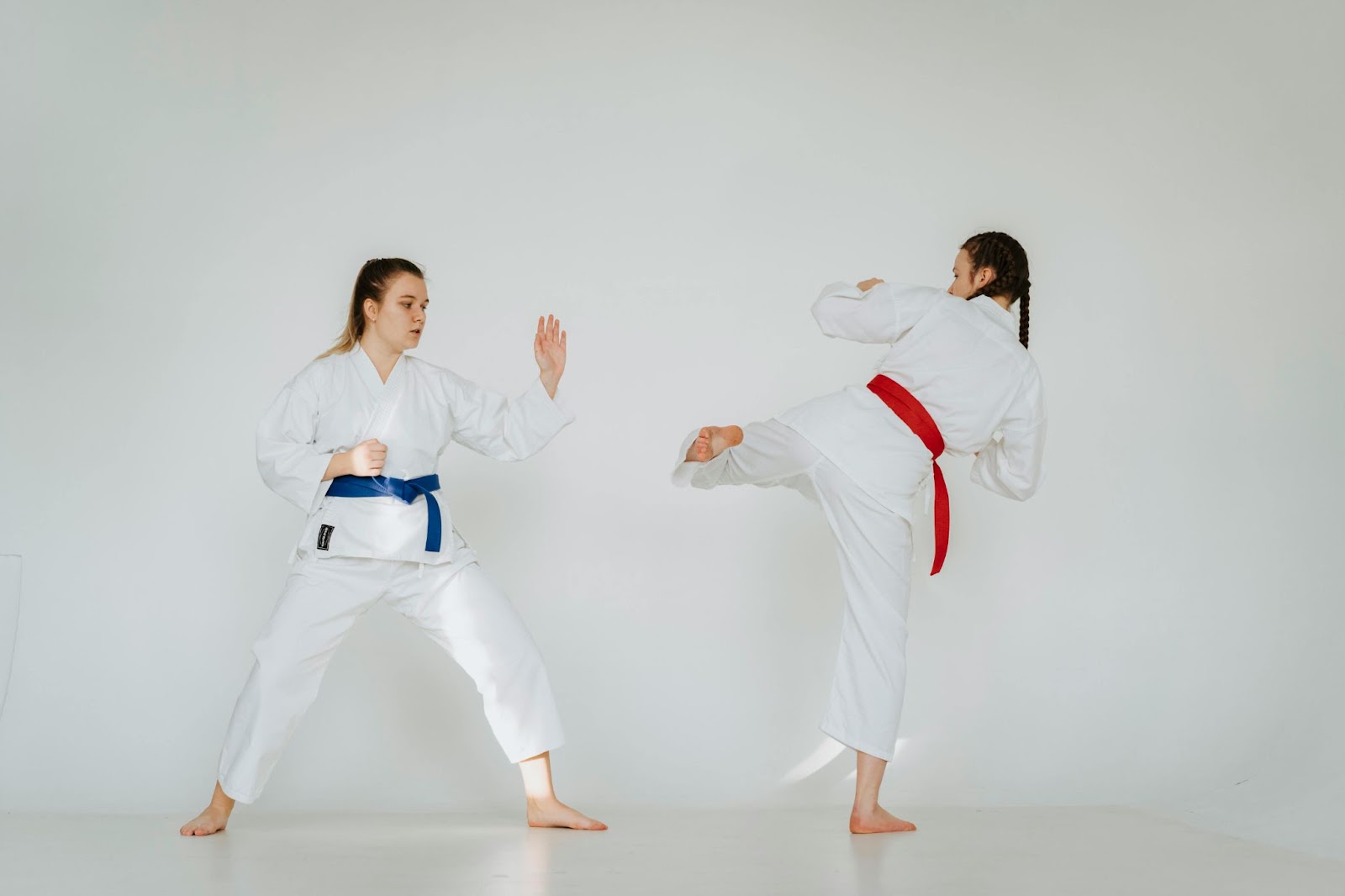 Two young female martial arts students practicing together. One student is mid-kick, while the other prepares to block with her arm.