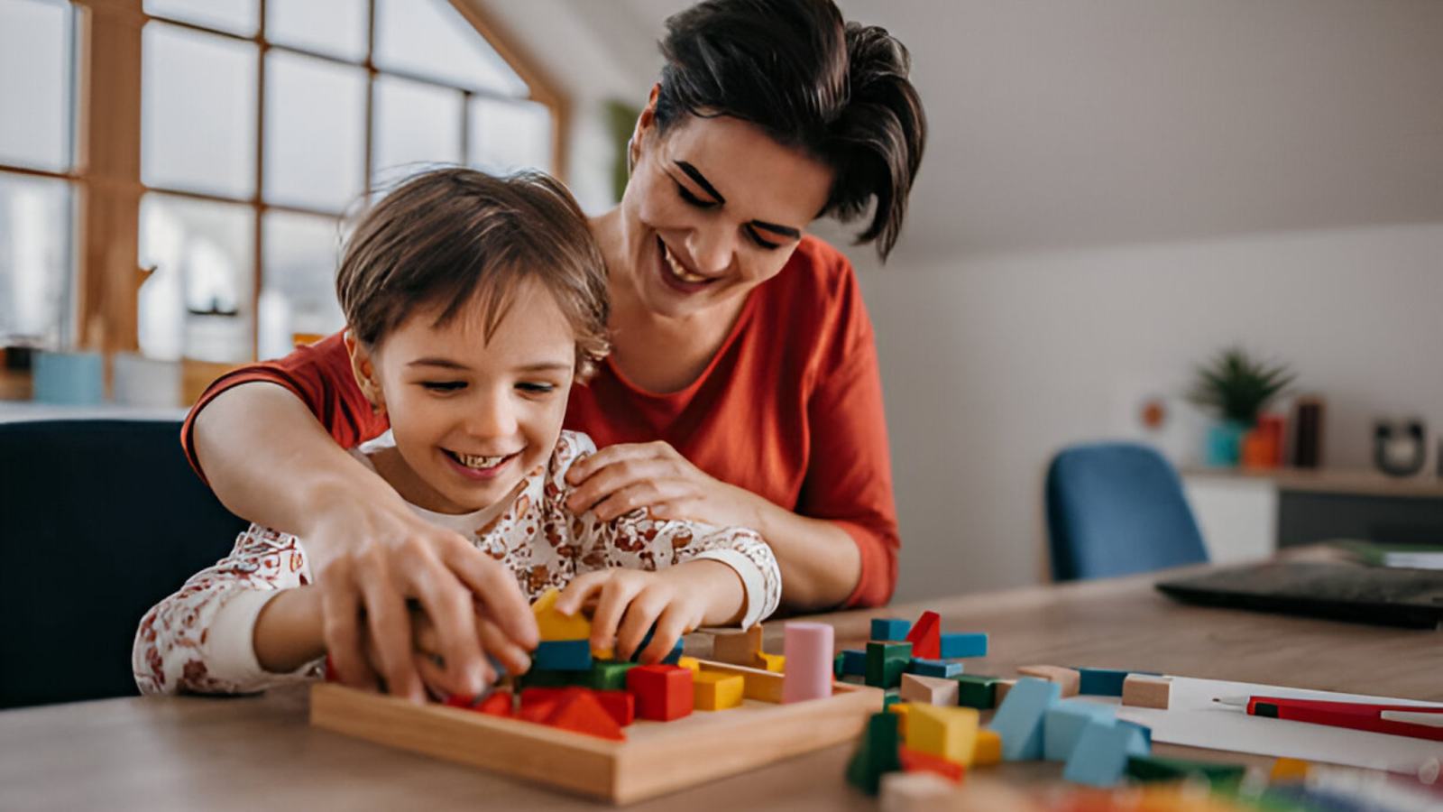 A kid and his mother are playing with building blocks. Colorful blocks are on the table
