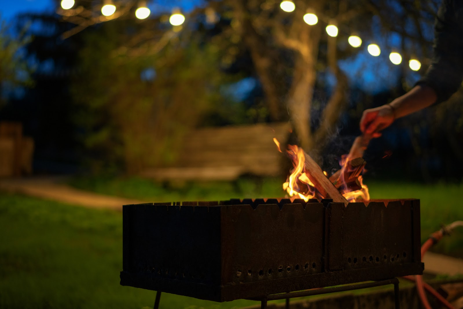 A cozy night garden scene illuminated by decorative lanterns hanging from the trees featuring wooden logs burning in a grill, casting a warm, inviting glow