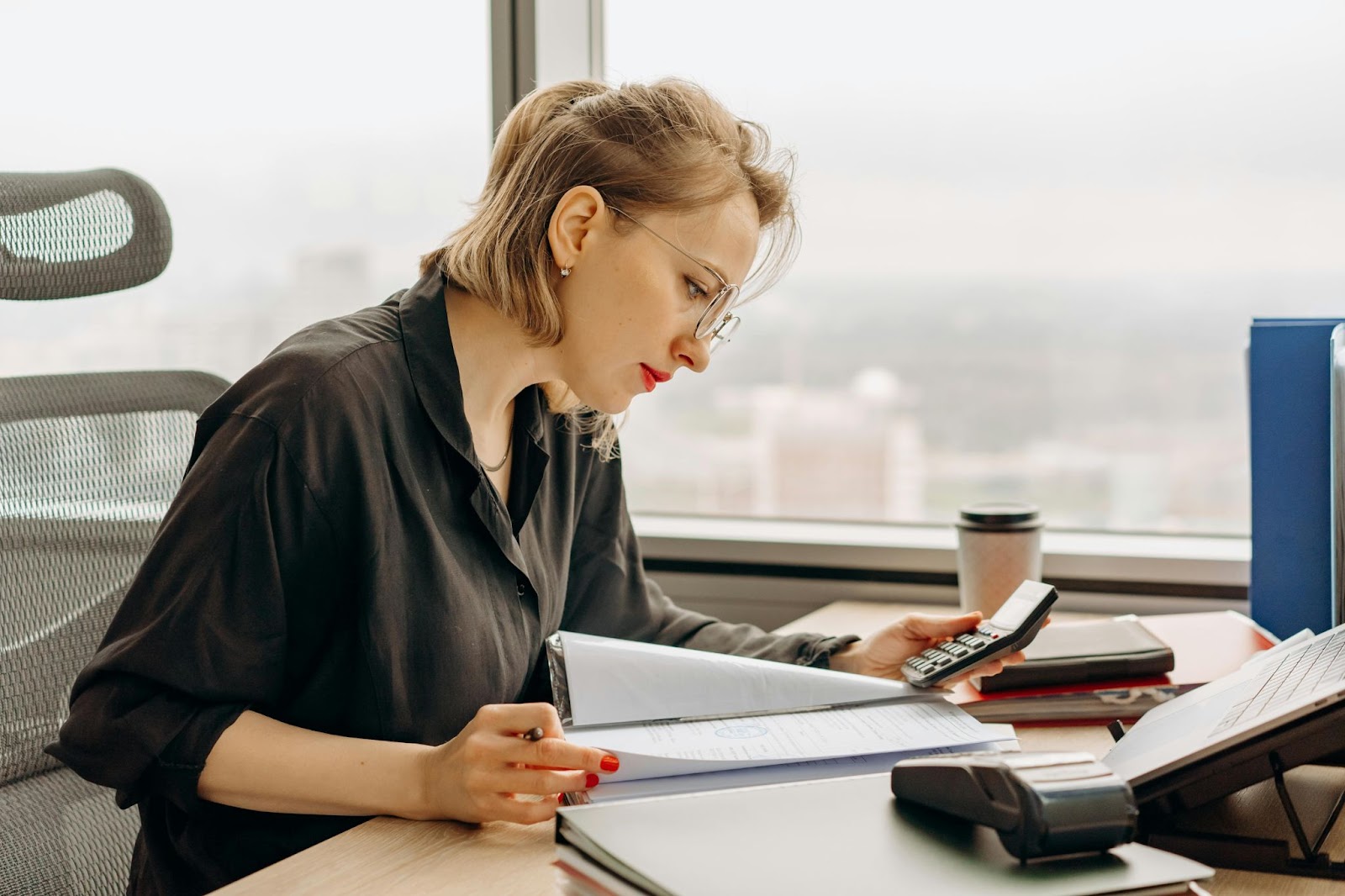 A woman looks over paperwork in an office while holding a pen and calculator.