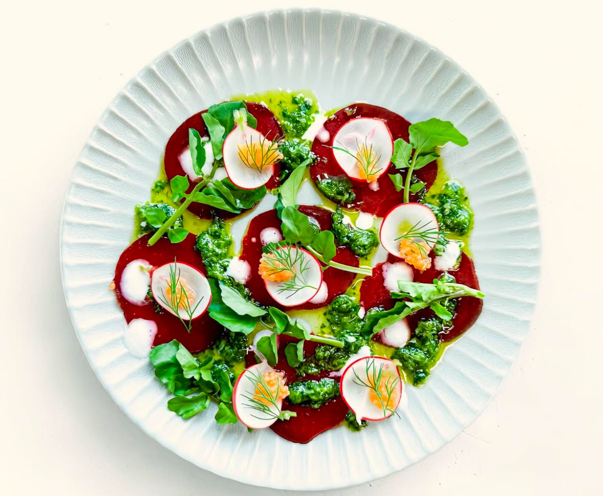 A beautifully plated dish arranged on a white, textured plate. The vibrant red beet slices are topped with thinly sliced radishes, dollops of creamy dressing, and fresh arugula leaves. Bright green pesto is drizzled throughout, adding a fresh and colorful contrast to the dish.&nbsp;&nbsp;