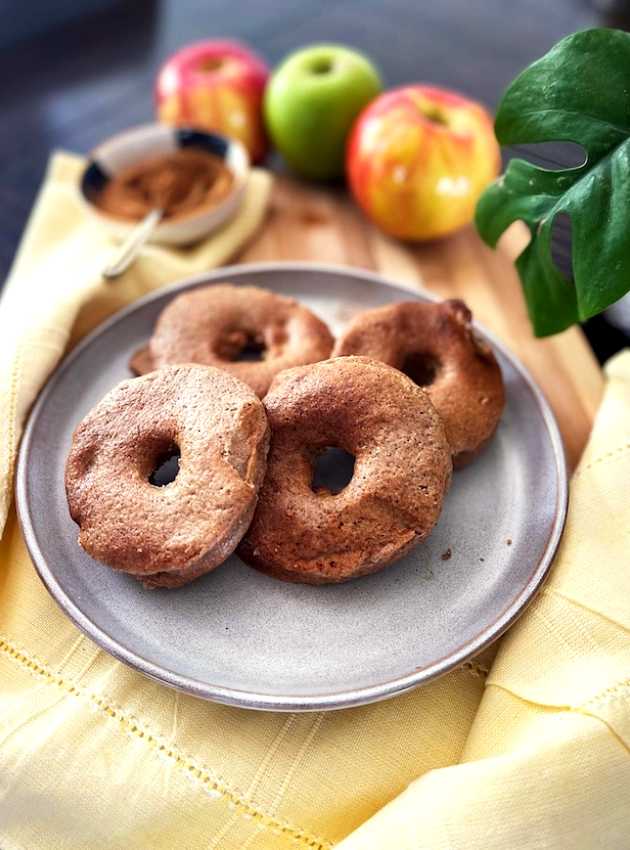 Four baked apple donuts on a plate with apples and cinnamon in the background.