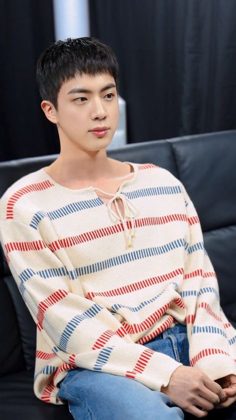 This contains a picture of BST Jin sitting on top of a black couch wearing a sweater and blue jeans