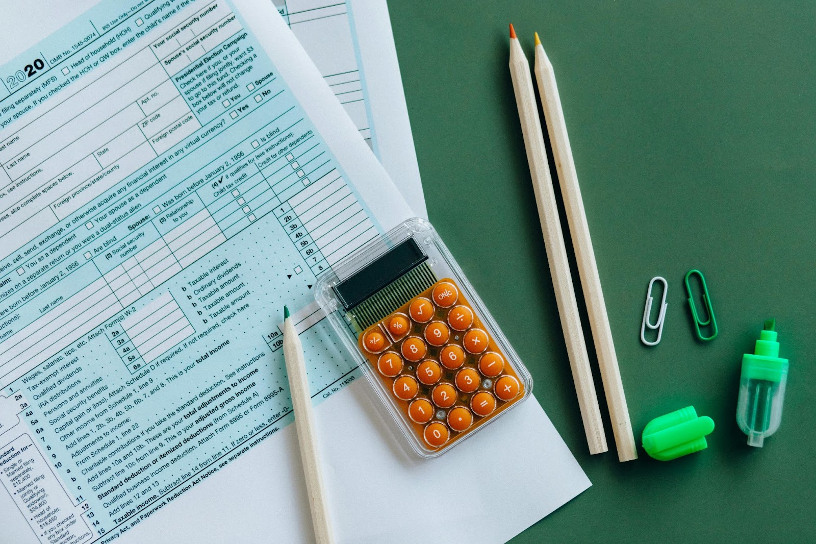 Una hoja de cálculo de impuestos junto a una calculadora naranja, lápices, clips y un borrador sobre un fondo verde