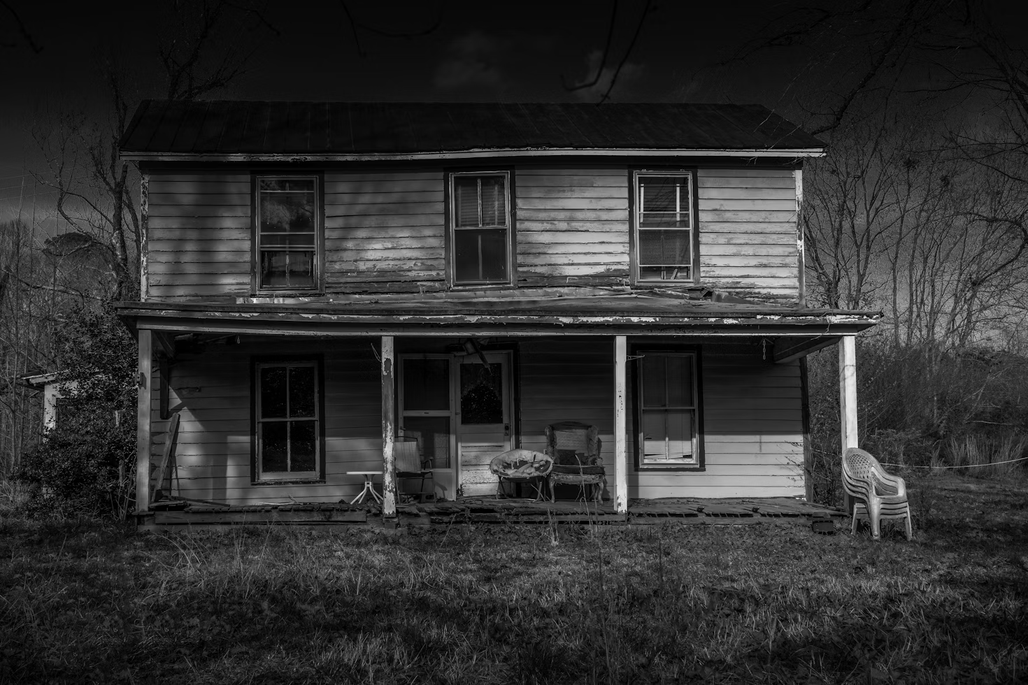 A rundown, abandoned two-story house with peeling paint, a dilapidated porch, and old furniture in front