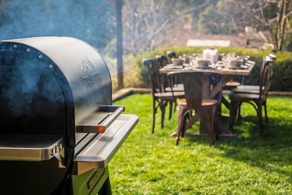 Traeger grill set in foreground with a picnic table in the background. Both on grass during the late afternoon sun.