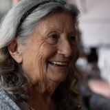 An elderly woman with long gray hair smiling warmly indoors.