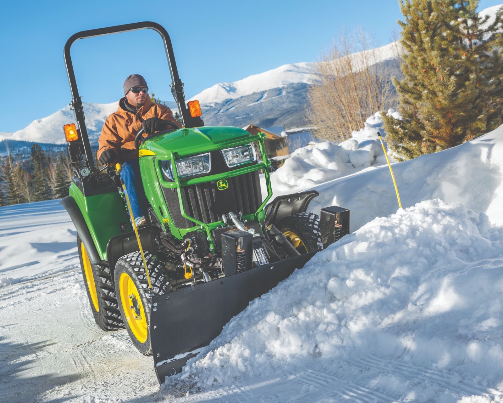 This image features a man operating a John Deere 2032R Compact Tractor equipped with a front blade attachment, clearing snow from a driveway or rural road in a winter landscape. The operator is wearing a brown winter jacket, gloves, sunglasses, and a beanie, suitable for cold weather conditions. The tractor is fitted with turf-friendly, deep-tread tires, likely for enhanced traction on snow and ice.  The background showcases a snow-covered mountainous region with pine trees and a clear blue sky, indicating a cold but sunny winter day. The bright yellow snow stakes on the side of the road help mark boundaries for snow removal.  The image conveys winter utility and efficiency, demonstrating the versatility of John Deere compact tractors for seasonal maintenance and snow management in residential, commercial, or rural settings.