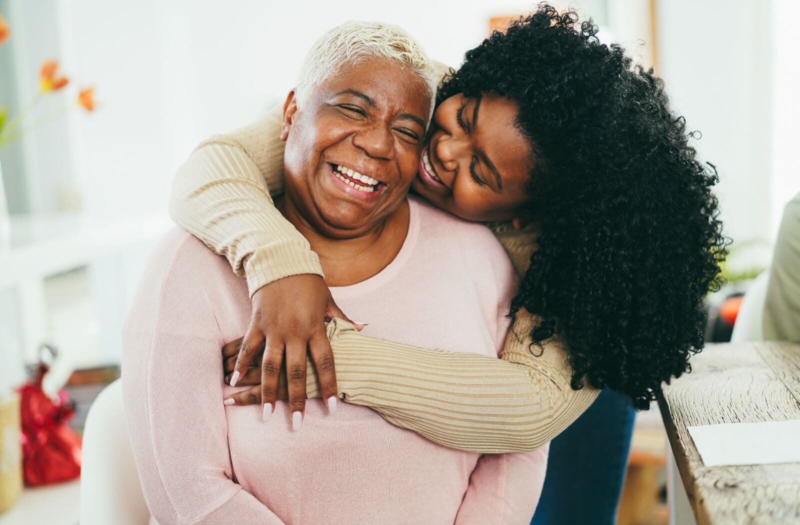 An adult child hugs their older parent from behind while visiting them in assisted living