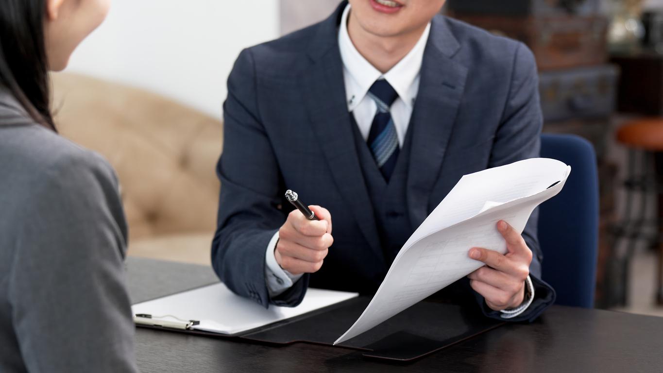 A man in a business suit holding a pen and papers while interviewing someone for a new job, going over an employee background check with the candidate.