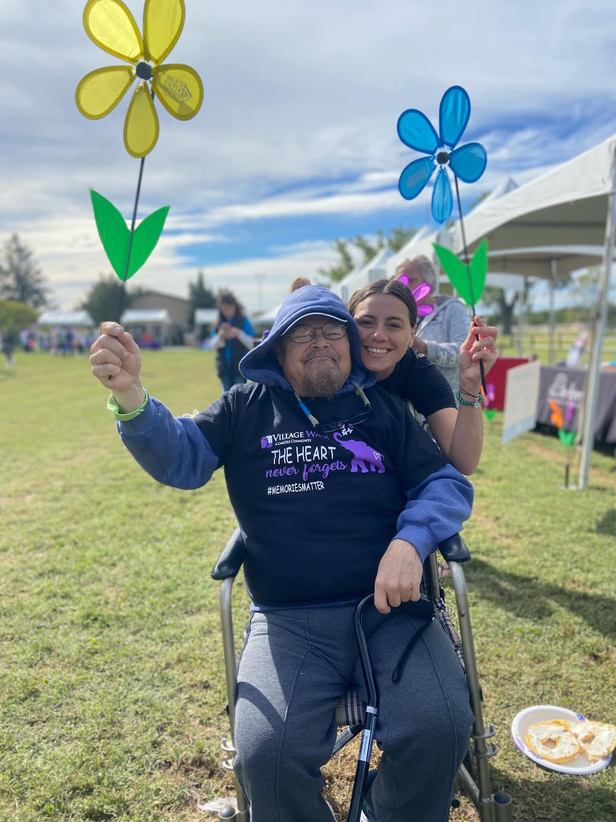 A senior citizen in a wheelchair  and his caregiver, both smiling and holding up plastic flowers