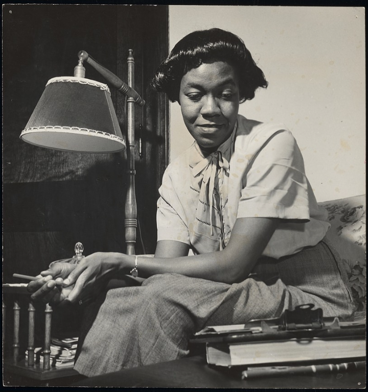 Image: Black-and-white photograph of Gwendolyn Brooks wearing a blouse and skirt while she rests her forearms on her lap, sitting on a floral chair next to a lamp, and glancing down at a clipboard with a pen in her hand. Photograph by Griffith Davis. Johnson Publishing Company Archive. Courtesy J. Paul Getty Trust and Smithsonian National Museum of African American History and Culture.