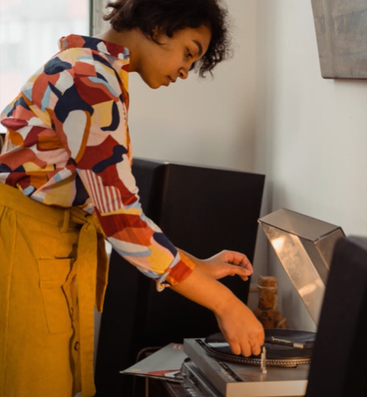 A person in a colorful shirt and yellow skirt adjusting a vinyl record on a turntable in a cozy room.