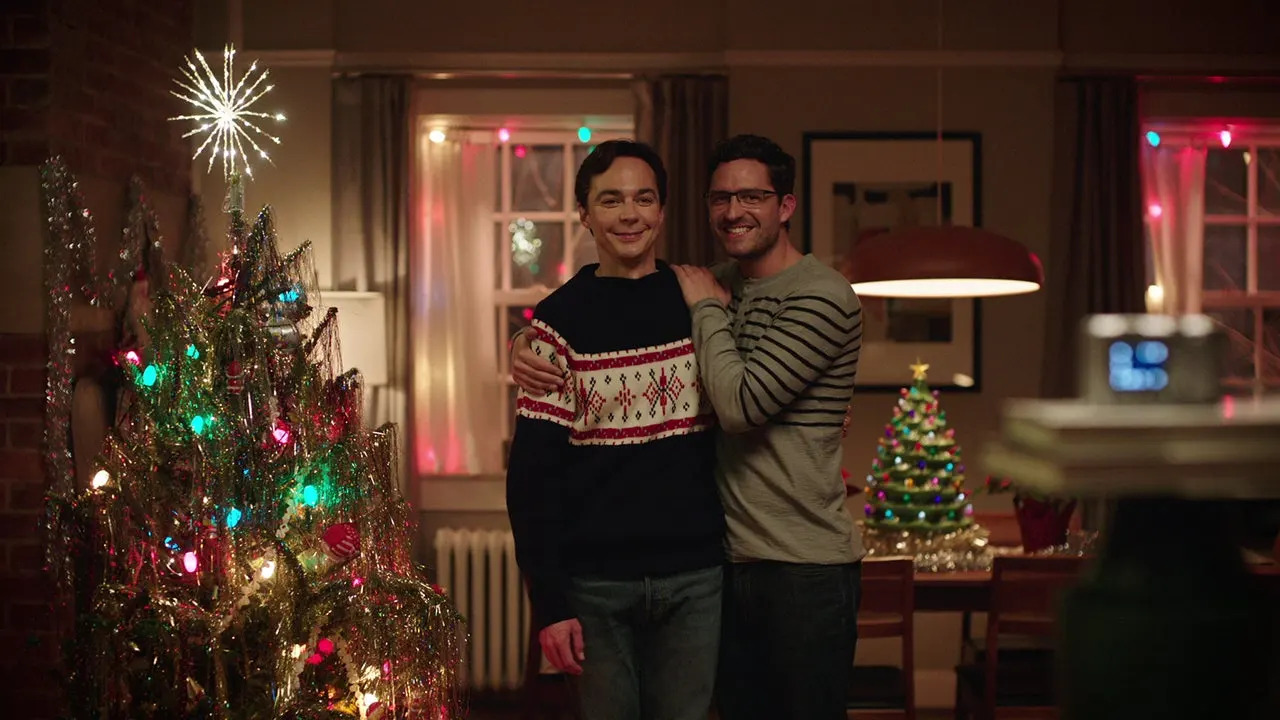 Pareja sonriente posando frente a un árbol de Navidad decorado, con luces navideñas en el fondo.