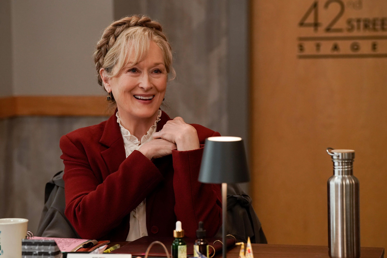 Meryl Streep sonriendo en una escena de la serie 'Only Murders in the Building', sentada en una mesa con accesorios, mostrando una actitud amigable y cálida.