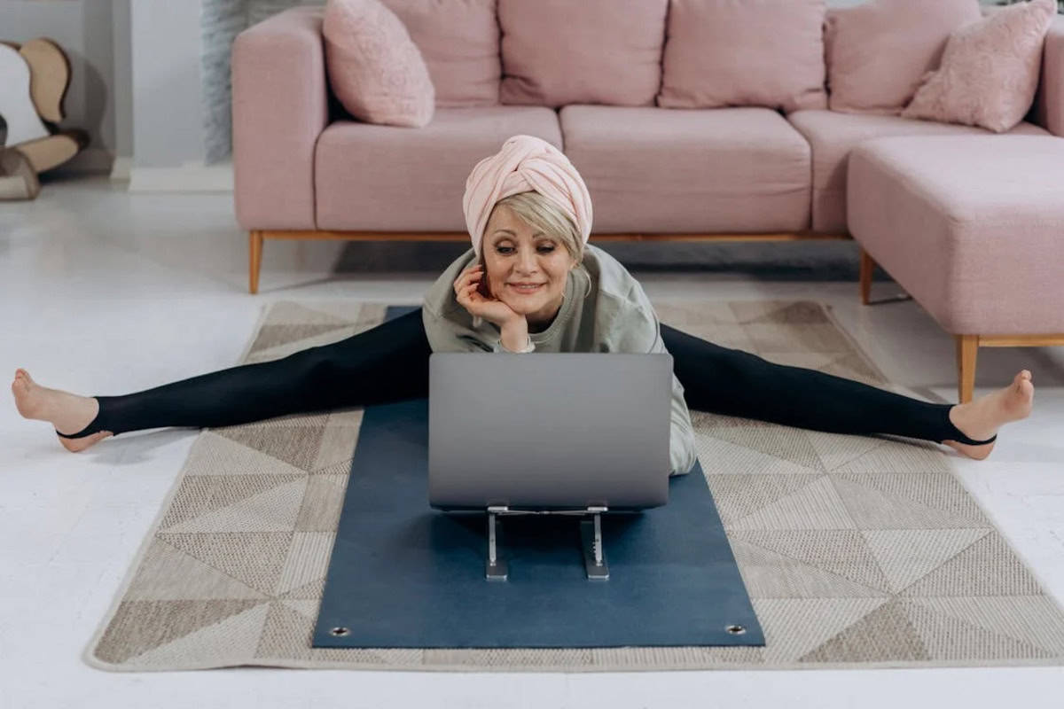 Mulher em uma sala praticando yoga, em postura de meditação, enquanto observa um notebook aberto, representando a prática de yoga online sendo o melhor estilo para iniciante.