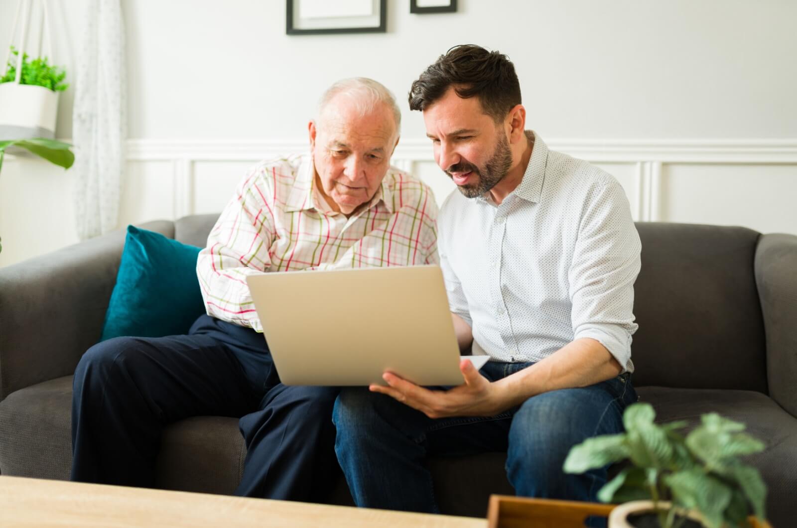 A family member sitting next to an older adult on a couch helping set up security measures on a laptop for online safety.
