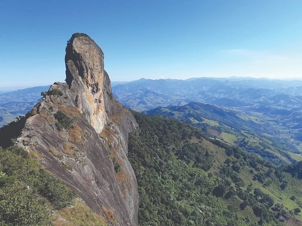 Paisagem de montanha com formação rochosa icônica na Serra da Mantiqueira, cercada por vegetação exuberante e vista panorâmica das colinas ao fundo, ideal para amantes de trekking e aventuras na natureza.
