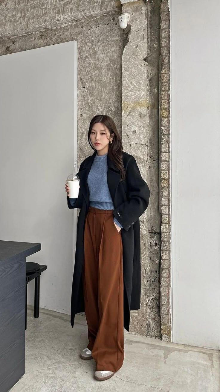 seorang wanita memakai celana cokelat, sweater biru, long coat hitam sedang memegang minuman