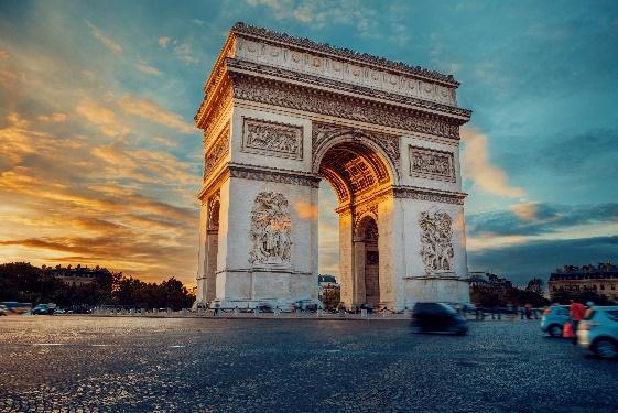 A large stone arch with a car driving on the road with Arc de Triomphe in the backgroundDescription automatically generated