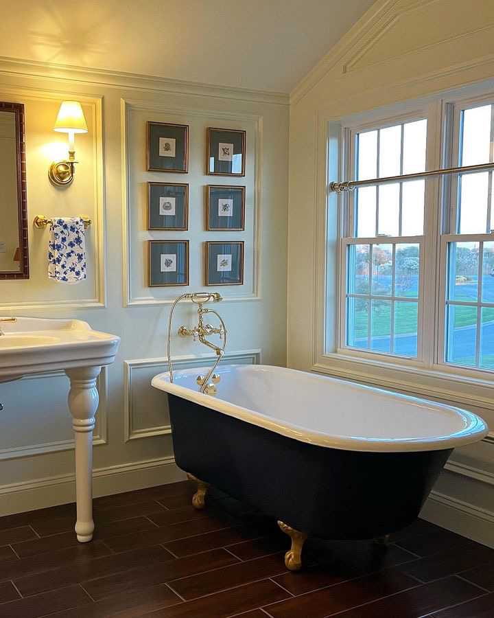 A black clawfoot tub with a brass faucet sits beneath six framed botanical prints on a wall. 