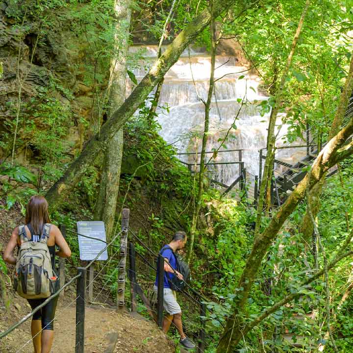 Casal de turistas explorando uma trilha na floresta com cachoeira ao fundo, cercados por árvores verdes e vegetação densa, em uma atividade de ecoturismo.