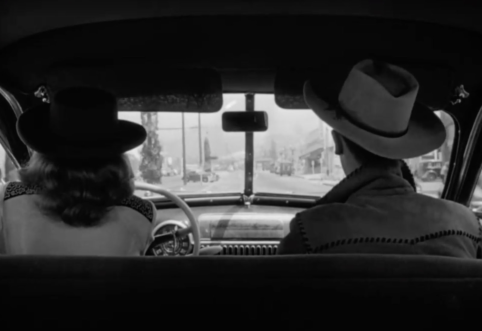 Silhouette of a woman driving, with a man seated on her right. The view out the windshield is a nondescript town.