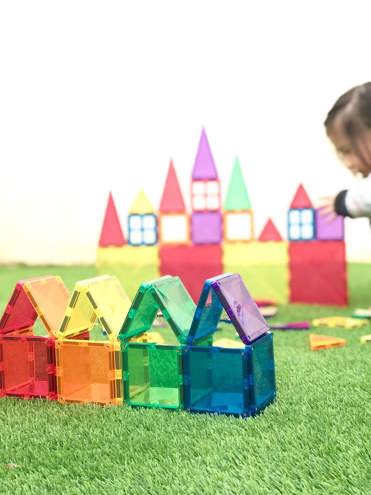 A toddler builds a castle with colourful magnetic tiles, including large and small squares, equilateral triangles, and isosceles triangles. Next to the castle is a row of rainbow-coloured houses made from the same vibrant tiles, neatly arranged in playful geometric shapes.