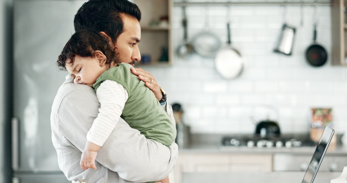 Delaware paid family leave: father carrying his sleeping baby while looking at a laptop
