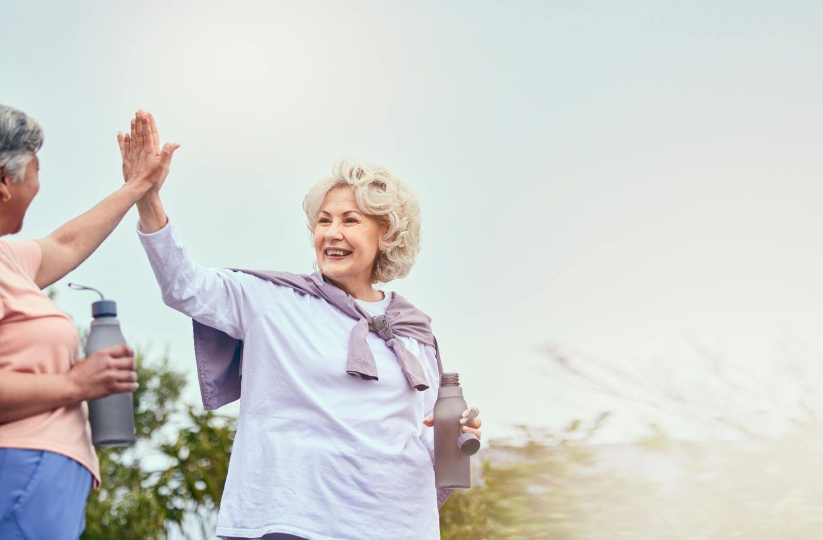 A pair of older adults holding water bottles and high-fiving while spending time outside in the park.