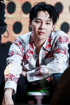 This contains an image of G Dragon  with black hair sitting down in front of a microphone and wearing a red and white shirt