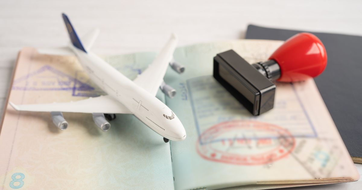 A miniature airplane model placed on an open passport with visa stamps, accompanied by a red stamp and a black ink pad. The scene symbolizes travel and visa processing.