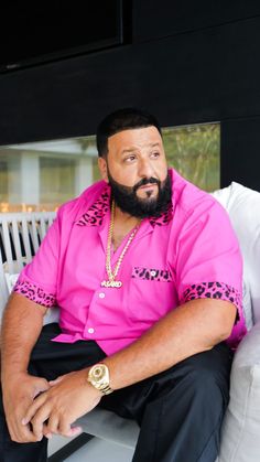 This contain DJ Khalid with a pink shirt and black pants sitting on a white couch