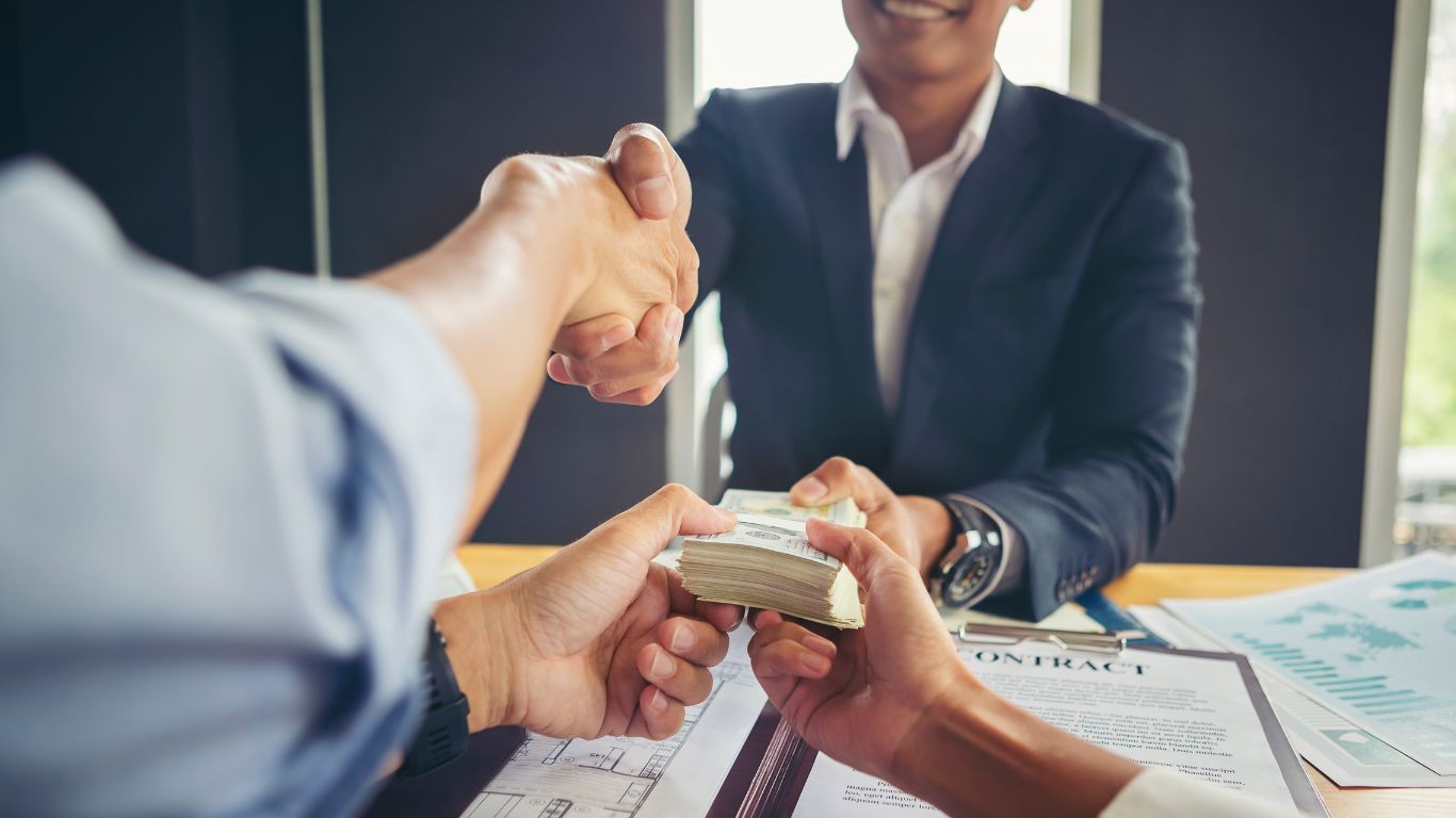 Two individuals shaking hands while exchanging a stack of money, with a contract and paperwork on the table, symbolizing a financial or business agreement.