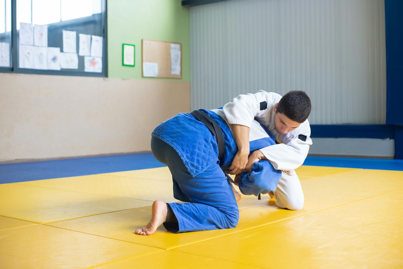 Two martial arts students kneeling on the floor, practicing headlock techniques during a training session in a dojo.






