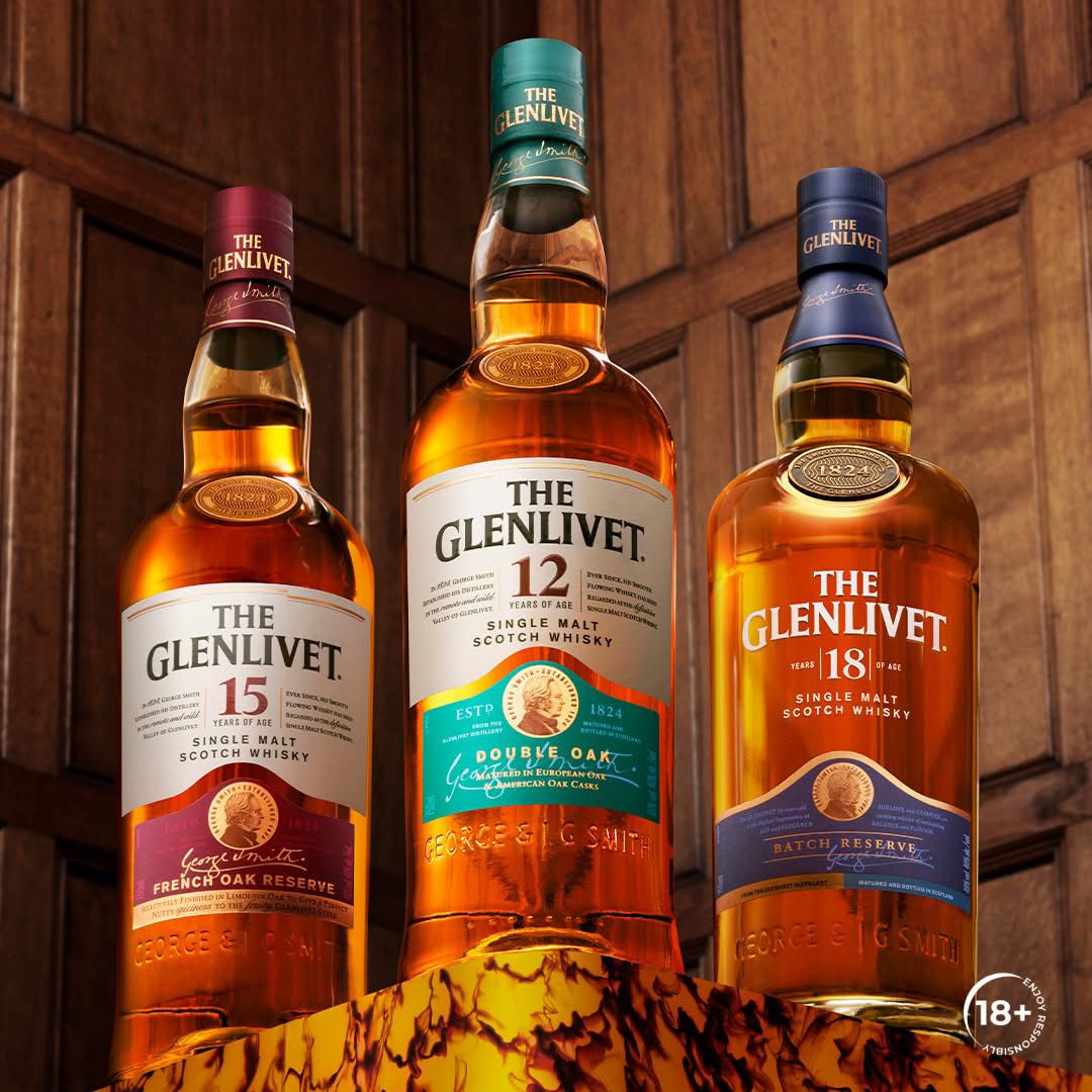 Three Glenlivet whisky bottles displayed on a wooden background, featuring Glenlivet 12, 15, and 18-year-old single malt options. The bottles are upright and well-lit, showcasing their labels and premium quality. 