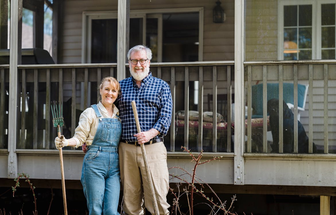 Free Elderly couple joyfully standing outside their home with gardening tools, symbolizing happiness and love. Stock Photo