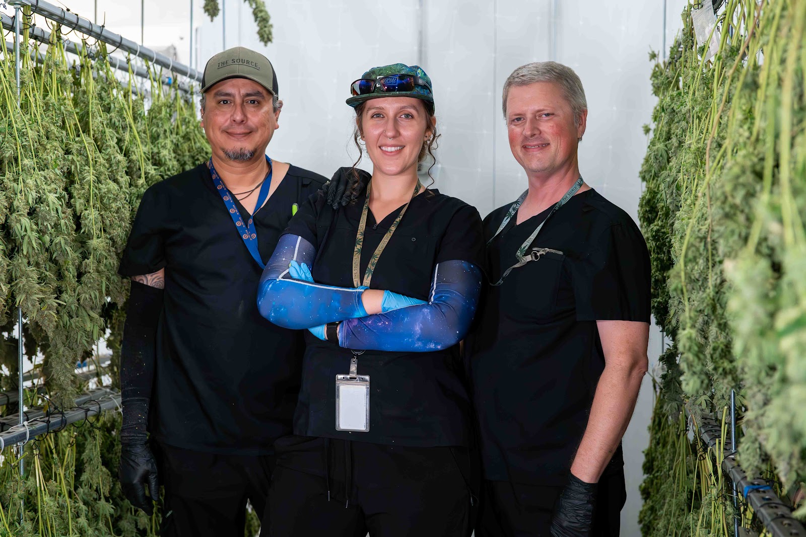 Portrait of three people from the waist up. A woman wearing a baseball cap stands in the middle with her arms crossed, and on both sides of her stand two men. All three people are smiling. In the foreground and background, marijuana is hanging to cure at The Source, a boutique cannabis grow, dispensary, and lab in Northwest Arkansas.