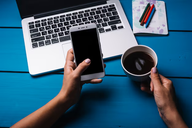 Shot of a person's hands holding a phone in one and a cup of coffee in the other in front of a laptop