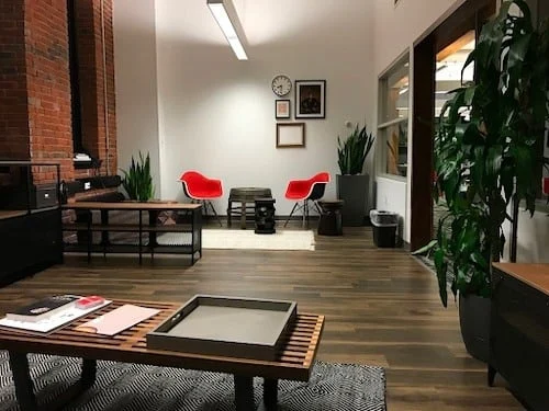 Modern office lounge with wooden floors, exposed brick, and contemporary furniture. Features red chairs, a coffee table, a wooden bench, and potted plants, with framed artwork and a clock on the wall.