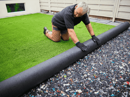 Worker installing eco friendly artificial grass in a residential backyard, unrolling turf over prepared ground.