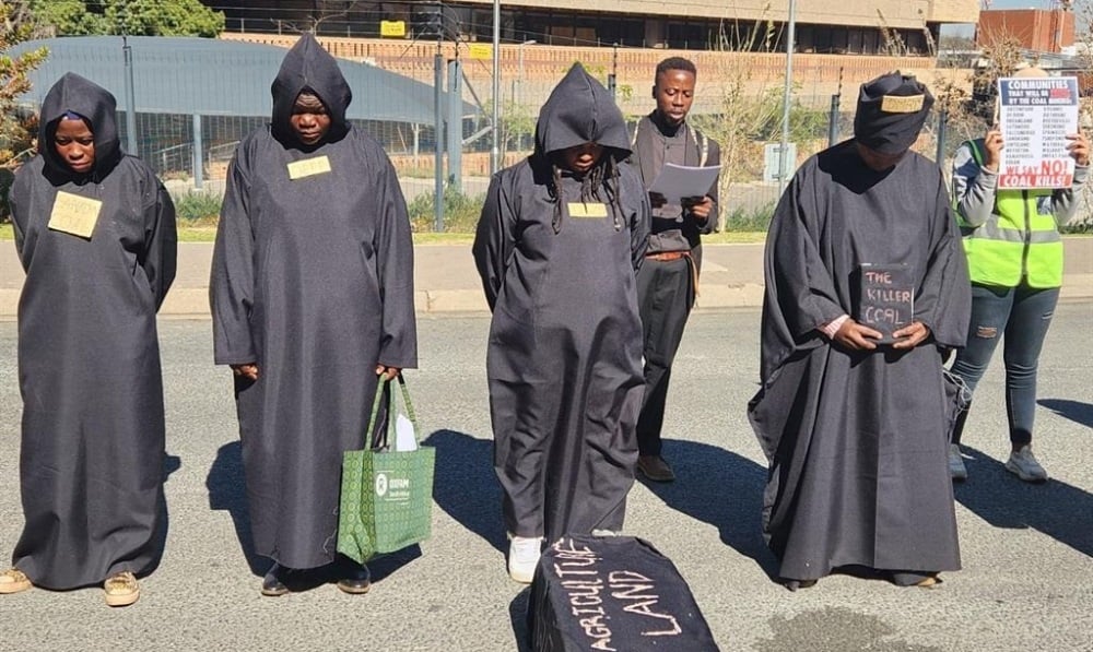 Rebels stand in a line, heads bowed, wearing black, before a coffin that says 'agriculture land'