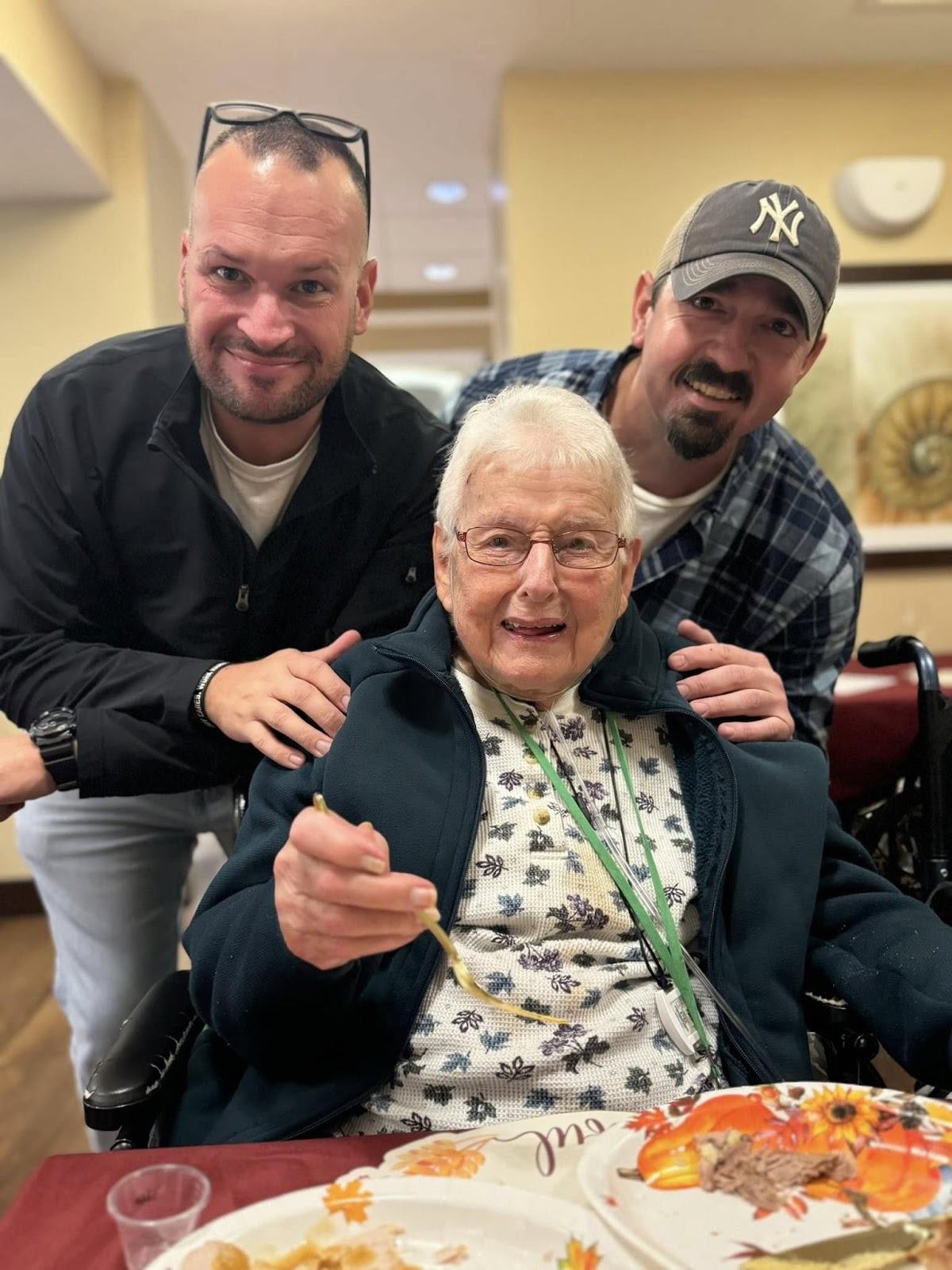 Two gentlemen smiling with their hands on the shoulders of a woman in a wheelchair in front of them.