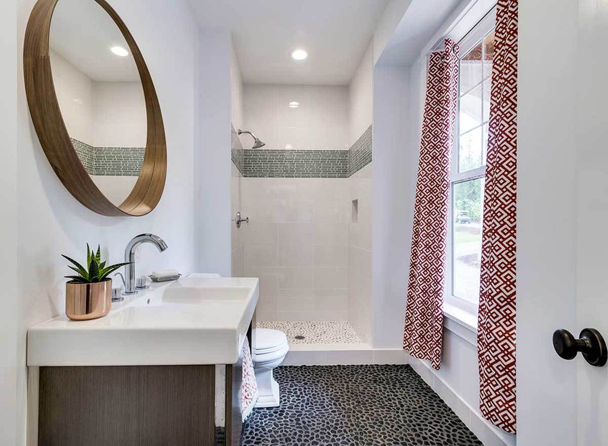 Bathroom with a sink vanity, a toilet, a walk-in shower, and a white framed window dressed in red patterned drapes.