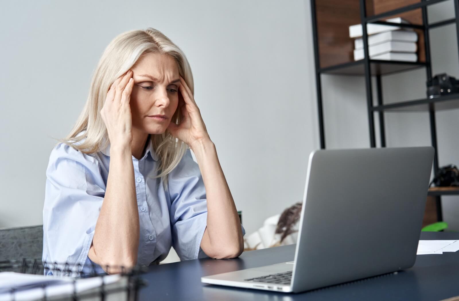 A middle-aged woman sits at her computer, looking frustrated and distressed and experiencing symptoms of early-onset dementia.