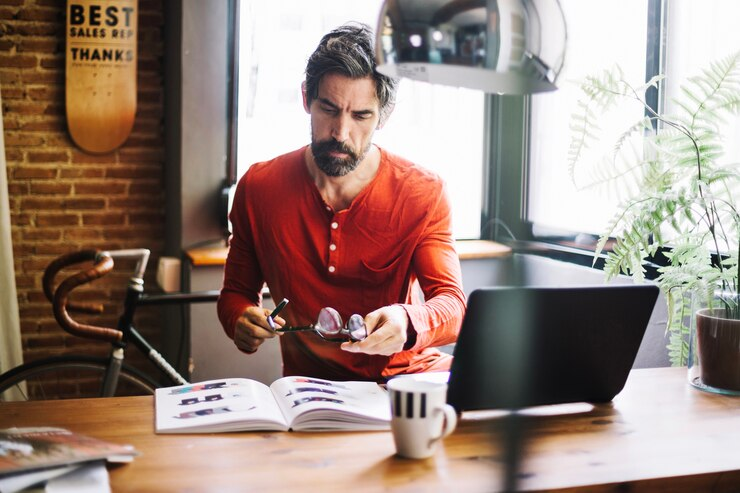 A stylish man reading a magazine while sitting in front of a laptop